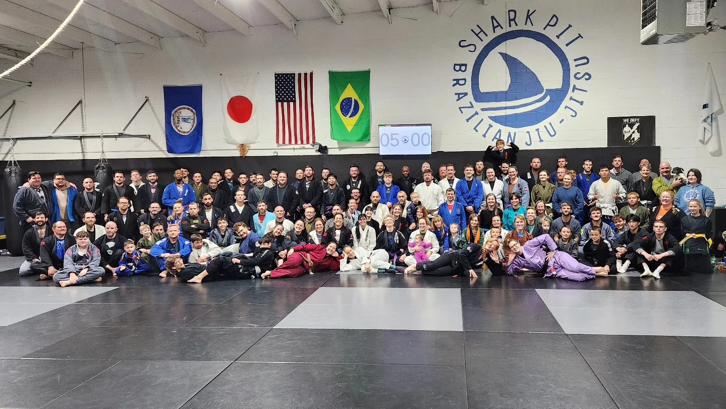 Large group of students and instructors posing on gym mats beneath the Shark Pit Brazilian Jiu-Jitsu logo and several flags
