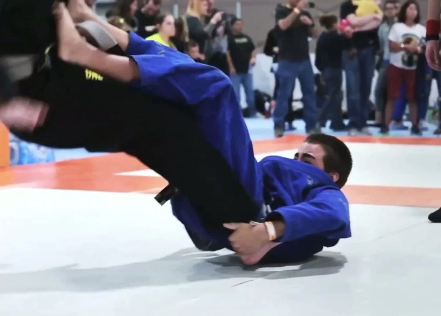 Competitor in a blue gi lies on his back beneath an opponent in black, with spectators watching at a tournament